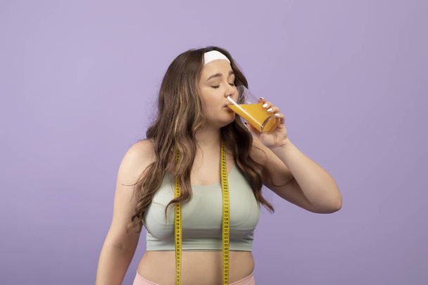 Calma bonita mujer joven de tamaño más en ropa deportiva con los ojos cerrados y cinta métrica beber vaso de jugo de naranja - Foto, imagen