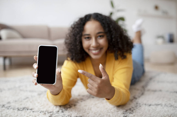 Vrolijke jonge Afro-Amerikaanse vrouw gericht op smartphone met leeg scherm, liggend op de vloer thuis, mockup - Foto, afbeelding