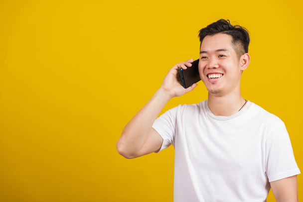 asiático guapo joven sonriendo positivo hablando en el teléfono celular, estilo de vida feliz macho usando móvil smartphone estudio disparo aislado en amarillo fondo - Foto, imagen
