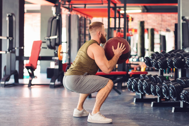 Un uomo atletico barbuto si accovaccia con una palla medica in palestra. Concetto di riscaldamento e fitness. - Foto, immagini