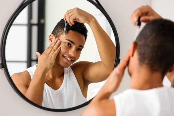 Jonge Afro-Amerikaanse man die serum gebruikt voor huidverzorging in de badkamer - Foto, afbeelding