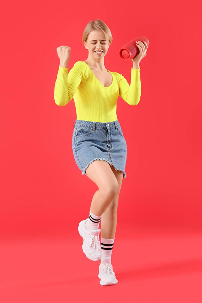 Young woman with modern wireless portable speaker dancing on red background - Foto, Bild