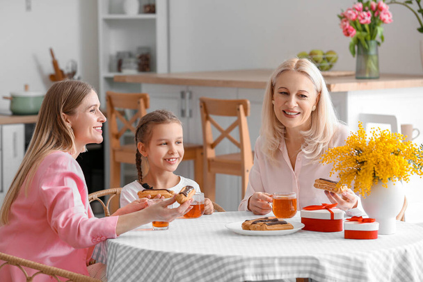 Młoda kobieta z córeczką i matką jedzą eklerki i piją herbatę w kuchni - Zdjęcie, obraz