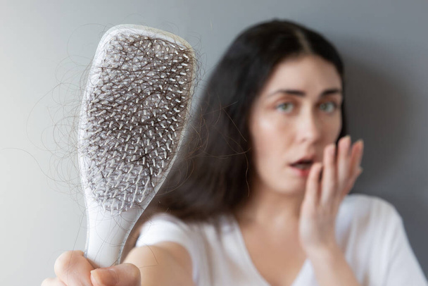 Blurred portrait of a shocked Caucasian young woman shows her hairbrush with losing hair. Gray background. The concept of baldness. - Valokuva, kuva