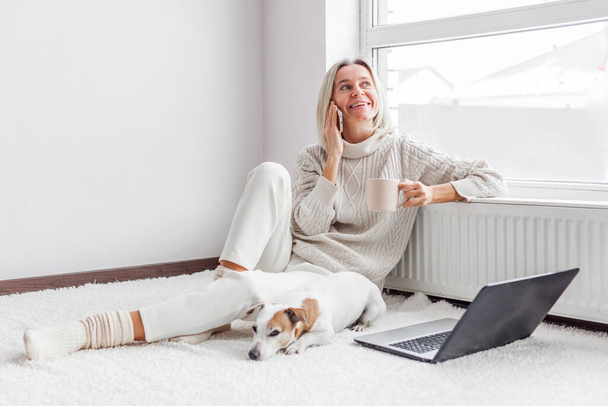 Happy middle aged woman with dog using her laptop and mobile phone at cozy white home. Female freelancer working in living roo - Fotografie, Obrázek