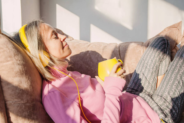 people, technology and leisure concept - happy middle-aged 40s woman sitting on sofa with headphones listening to music at home - 写真・画像