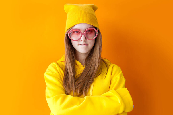 Modern confident teen girl in trendy glasses wearing yellow hoodie with knitted hat. Young millennial woman looking at camera on orange background - Фото, изображение