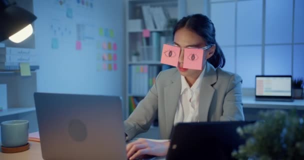 Masada oturan Asyalı genç iş kadını geceleri ofiste dizüstü bilgisayar başında uyuyakalıyor. Yorgun bir kadın. Gözlerinin üzerinde iki post-it var. İş yerinde uyuyan yüz üzerine yapışkan notlar.. - Video, Çekim