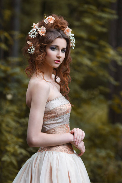 A slender girl in a long light dress with flowers in her hairstyle outside. - Foto, Imagem