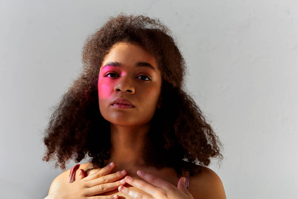 Δημιουργικό πορτρέτο στούντιο του νεαρού σγουρά αφρικανικό αμερικανικό μοντέλο με φωτεινό χρώμα art μακιγιάζ κοιτάζοντας κάμερα, μαύρη γυναίκα με ροζ χρώμα στο ένα μισό του προσώπου ποζάρουν απομονώνονται σε γκρι φόντο - Φωτογραφία, εικόνα