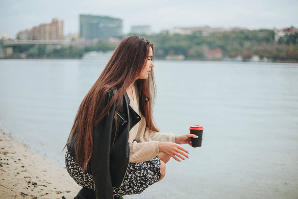 Mladá žena s odnést kávu v ruce v ležérní oblečení dřepí v blízkosti řeky vody na podzim den s městskou krajinou na pozadí, romantické snění žena na pobřeží těší rekreaci v přírodě - Fotografie, Obrázek