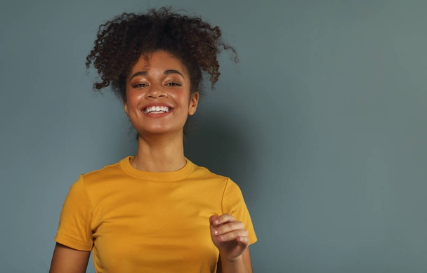 Όμορφη ευτυχισμένη σκουρόχρωμη αφροαμερικάνικη εθνικότητα γυναίκα με κίτρινο μπλουζάκι που σηκώνει το χέρι χαιρετώντας ενώ κοιτάζει στην κάμερα με ευχάριστο χαμόγελο, ποζάροντας ενάντια στο γκρι πράσινο φόντο του στούντιο - Φωτογραφία, εικόνα