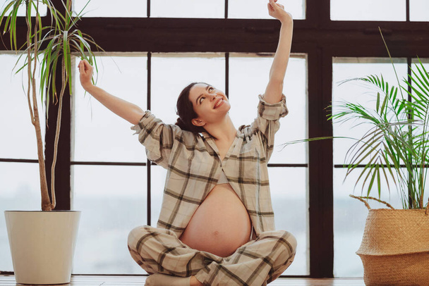 Longitud completa de la joven tranquila mujer embarazada en pijama sosteniendo el vientre mientras se sienta en pose de loto contra la ventana panorámica, la futura madre tocando suavemente la barriga. Meditación durante el embarazo - Foto, Imagen