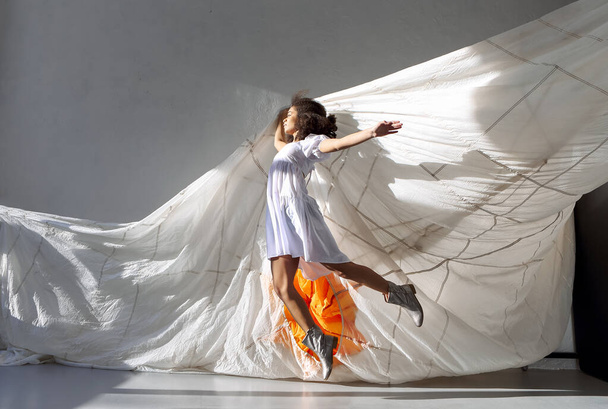 Jump and fly. Young overjoyed carefree african american woman jumping in air with big white parachute isolated over grey studio background. Freedom and thirst for life concept - Φωτογραφία, εικόνα