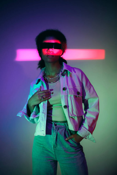 Young confident african american woman wearing VR headset exploring virtual world while standing in denim casual clothes under neon light in studio, playing virtual reality games. Modern technologies - Foto, afbeelding