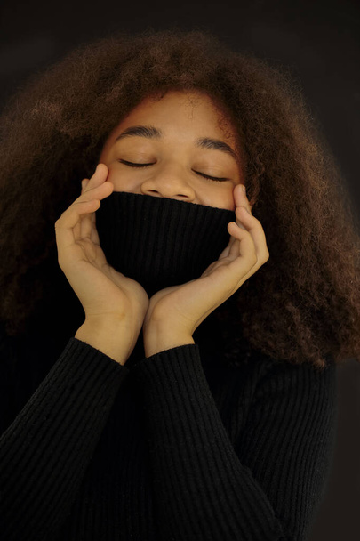 Pleasant Afrikaans amerikaanse krullende vrouw sluit de ogen en bedekt gezicht met warme zwarte trui kraag geïsoleerd op donkere studio achtergrond, glimlachende vrouw opwarmen in polo nek trui. Verticale foto - Foto, afbeelding