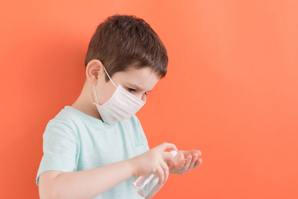 Preschool caucasian boy in white medical mask with sanitizer bottle on orange background. Coronavirus prevention concept - Foto, Bild