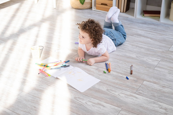 lockiges Kindermädchen zeichnet mit farbigen Filzstiften, die im heimischen Wohnzimmer auf dem Boden liegen - Foto, Bild