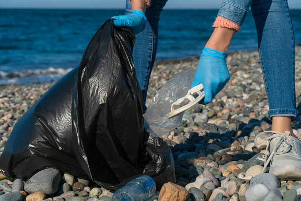 Gros plan des mains d'une femme qui ramasse des déchets en plastique et en caoutchouc dans un sac à ordures noir sur une plage de galets de la mer. Pollution de l'environnement sur la côte de la mer Noire. - Photo, image