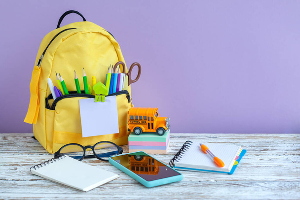 Concept back to school. Full yellow school backpack and toy bus school with stationery on table on purple background. - Zdjęcie, obraz