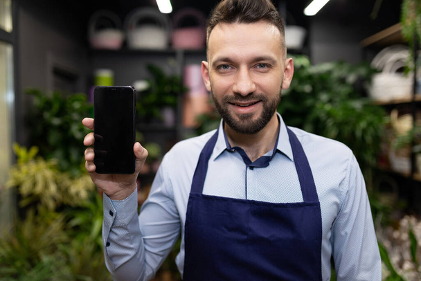 een bloemverkoper tegen de achtergrond van een bloemist winkel demonstreert een smartphone scherm met een glimlach op zijn gezicht - Foto, afbeelding