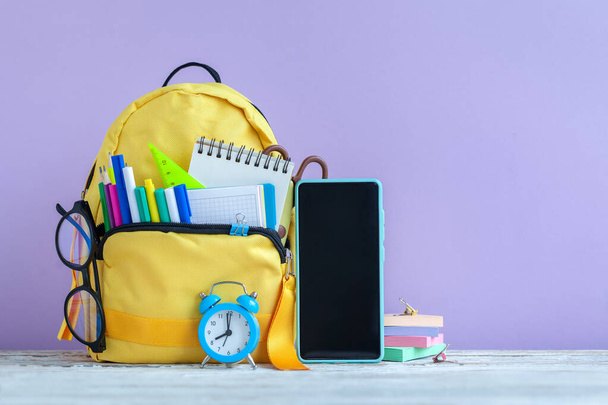 Concept back to school. Full yellow school backpack and smartphone, eyeglasses, alarm clock with stationery on table on purple background. - Fotó, kép