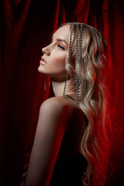 Krásná blondýnka s dlouhými vlasy na červeném sametovém pozadí. Šperky na hlavě ženy, moderní make-up a šipky na očích - Fotografie, Obrázek