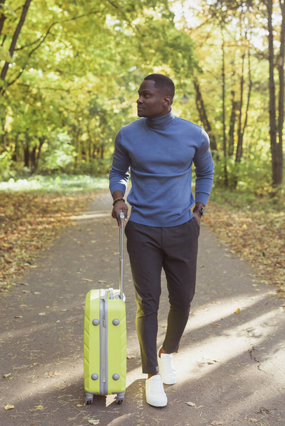 Cheerful african american young man in stylish clothes with suitcase walks in an spring park on warm sunny day. Countryside travel concept - Photo, Image