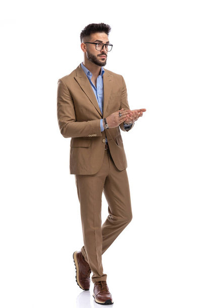 молодий бізнесмен в коричневому костюмі в окулярах дивиться в бік і натирає долоні під час ходьби ізольовано на білому тлі в студії
 - Фото, зображення