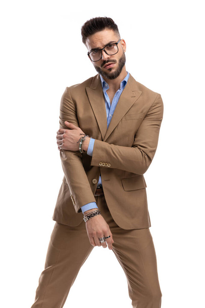 homme d'affaires sexy non rasé avec des lunettes tenant le coude dans une pose de mode tout en se tenant debout et posant isolé sur fond blanc en studio - Photo, image