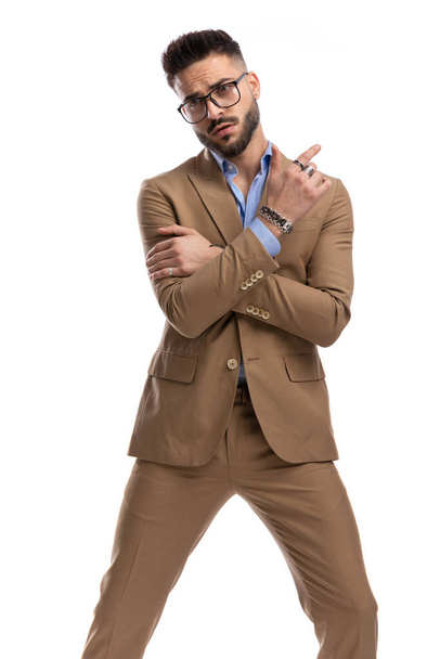 sexy junger Kerl im braunen Anzug mit Brille, die Arme verschränkt und mit dem Finger nach hinten zeigt auf weißem Hintergrund im Studio - Foto, Bild