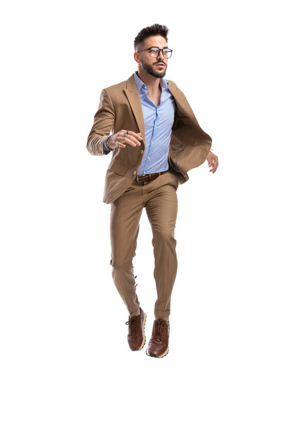 unshaved young guy in brown suit with glasses jumping and looking to side on white background in studio - Photo, image