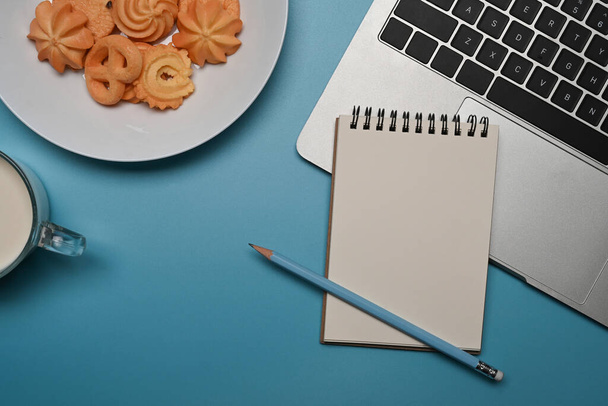 Тарелка с датским маслом печенье, свежее молоко, ноутбук и ноутбук на синем фоне. - Фото, изображение