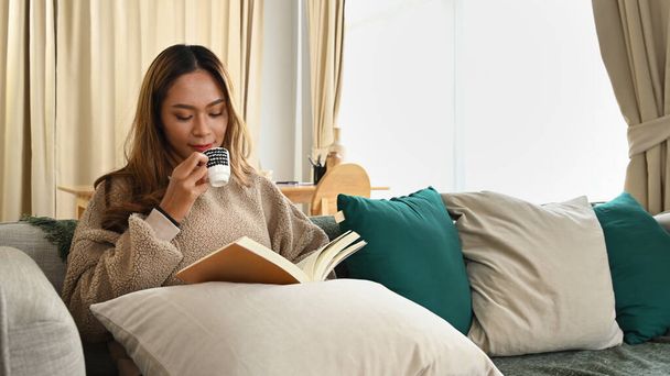 Мирная азиатская женщина читает книги и пьет кофе на диване. - Фото, изображение