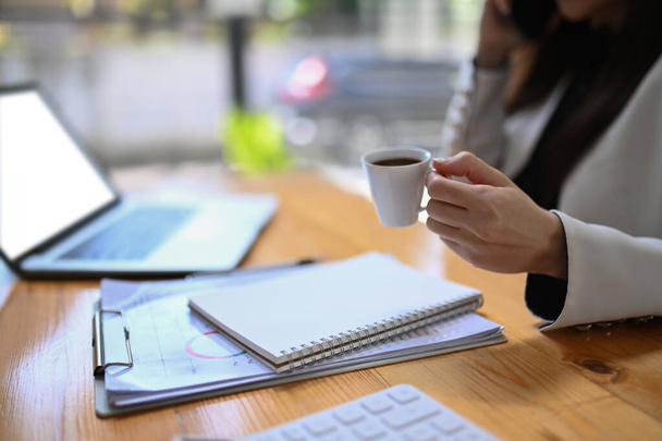 Обрезанный снимок деловая женщина держит чашку кофе и работает с ноутбуком в офисе. - Фото, изображение