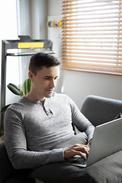 笑顔若い男がソファに横たわって、ラップトップコンピュータでインターネットを閲覧. - 写真・画像