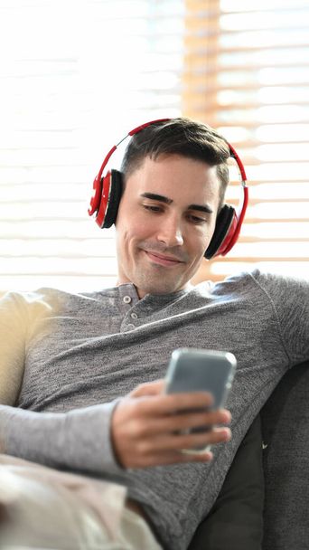 Portrait of handsome man wearing wireless headphone and using smart phone on sofa. - Фото, изображение