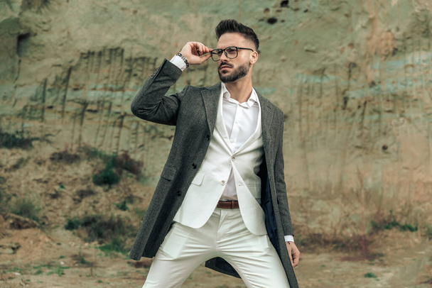 portrait of bearded young businessman looking to side and adjusting eyeglasses while posing outside in nature - Photo, Image