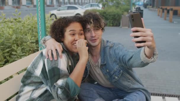 Mediana larga de estudiante universitaria negra sonriendo, charlando y tomando selfie en un teléfono inteligente con un amigo caucásico, sentada en el columpio en la zona peatonal en la fría noche de verano - Metraje, vídeo