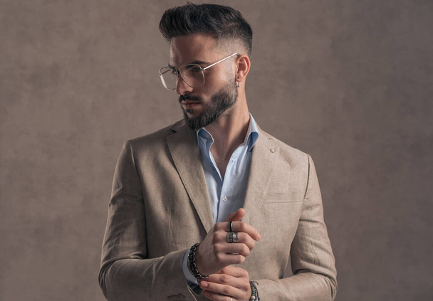portrait of bearded young man with eyeglasses looking to side and fixing sleeve on brown background in studio - Foto, immagini