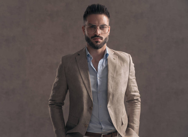 joven guapo con barba y gafas cogidas de la mano en los bolsillos y posando delante de fondo marrón en el estudio - Foto, Imagen