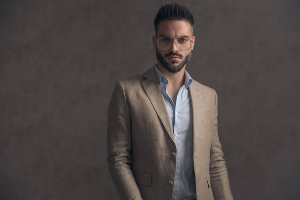 portrait of sexy unshaved man with glasses in beige suit posing in a cool manner on brown background in studio - Φωτογραφία, εικόνα