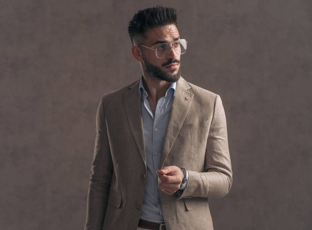 sexy barbudo hombre de negocios con gafas en traje beige mirando a un lado y posando sobre fondo marrón en estudio - Foto, imagen