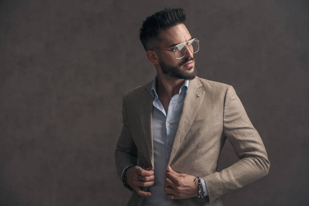 portrét pohledného mladého podnikatele s brýlemi rozhlížejícími se na stranu, zapínání a zavírání obleku na hnědém pozadí ve studiu - Fotografie, Obrázek