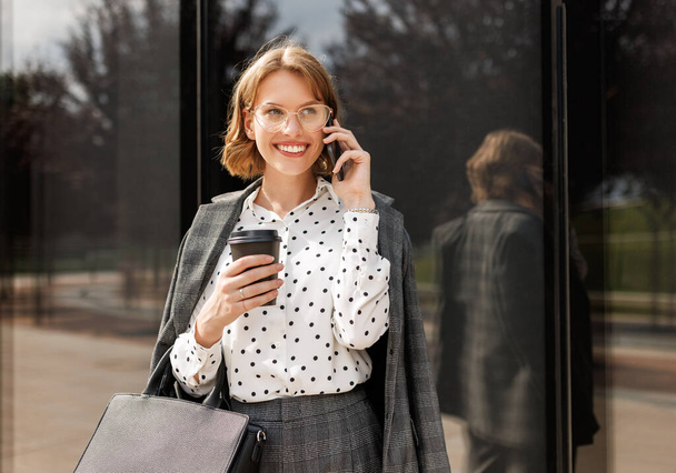 Senhora de negócios atraente fazendo chamada enquanto caminhava ao longo da rua da cidade com tomar café na mão - Foto, Imagem