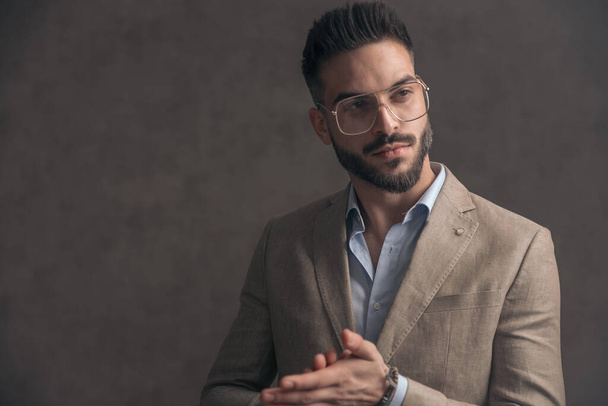 sexy young man with eyeglasses in beige suit looking to side and rubbing palms on brown background in studio - Foto, Imagem