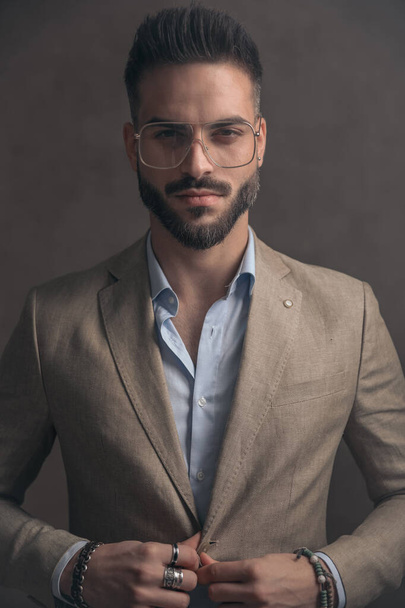 feliz joven hombre de negocios con gafas de cierre y ajuste de traje mientras posando sobre fondo marrón en el estudio - Foto, Imagen