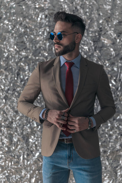 vonzó szakállas férfi retro napszemüveg keres oldalán és kigomboló kabát előtt ezüst alufólia háttér stúdióban - Fotó, kép