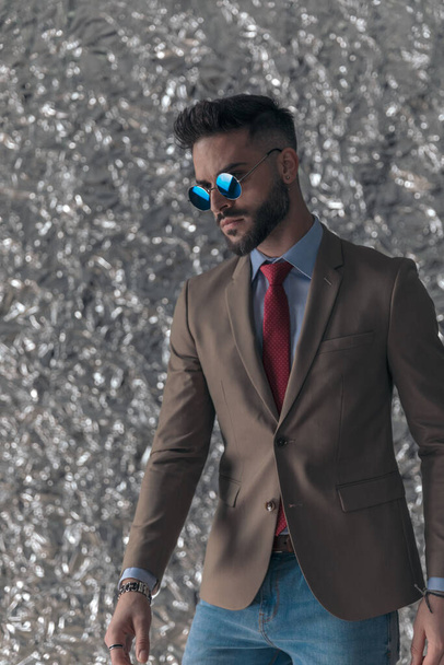 bearded young man with retro sunglasses looking to side and posing in front of silver tinfoil background - Фото, изображение