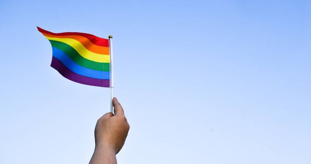 Orgullo arco iris lgbt banderas gay sosteniendo en la mano y siendo ondeado en la brisa contra el cielo nublado y azul. Lgbtq + celebra en concepto de mes de orgullo, enfoque suave y selectivo en banderas. - Foto, imagen